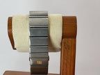 A stainless steel link Omega Constellation watch band with a gold buckle clasp, displayed on a cream-colored velvet watch stand set against a wooden background.