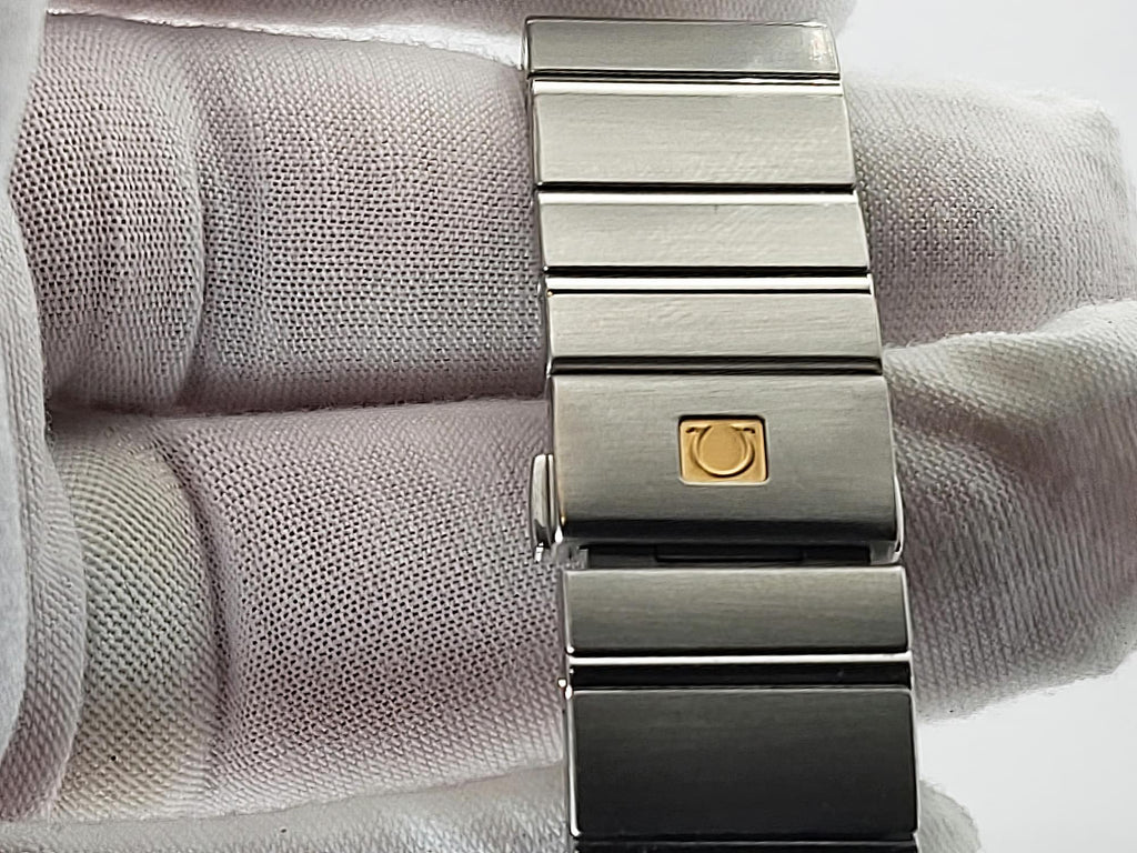 A close-up of a stainless steel Omega Constellation watch band clasp with a gold-tone circular logo, shown resting on a white textured fabric surface.