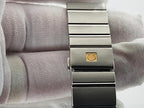 A close-up of a stainless steel Omega Constellation watch band clasp with a gold-tone circular logo, shown resting on a white textured fabric surface.