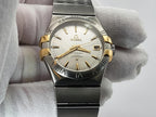 An Omega Constellation wristwatch with a two-tone stainless steel and gold-tone case, held by a hand wearing a white textured glove. The watch features a white dial with gold-tone hands, markers, and a date window.