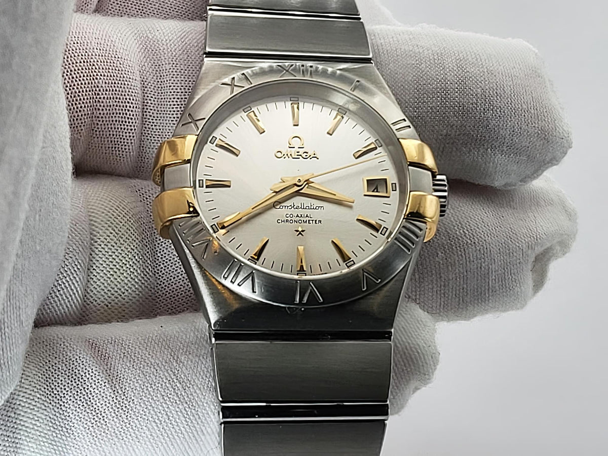 An Omega Constellation wristwatch with a two-tone stainless steel and gold-tone case, held by a hand wearing a white textured glove. The watch features a white dial with gold-tone hands, markers, and a date window.