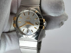 A two-tone Omega Constellation watch with a silver stainless steel case and bracelet, gold-tone hands and markers, and a white dial, held by a white textured glove.