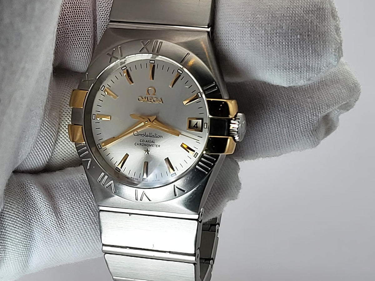 A two-tone Omega Constellation watch with a silver stainless steel case and bracelet, gold-tone hands and markers, and a white dial, held by a white textured glove.