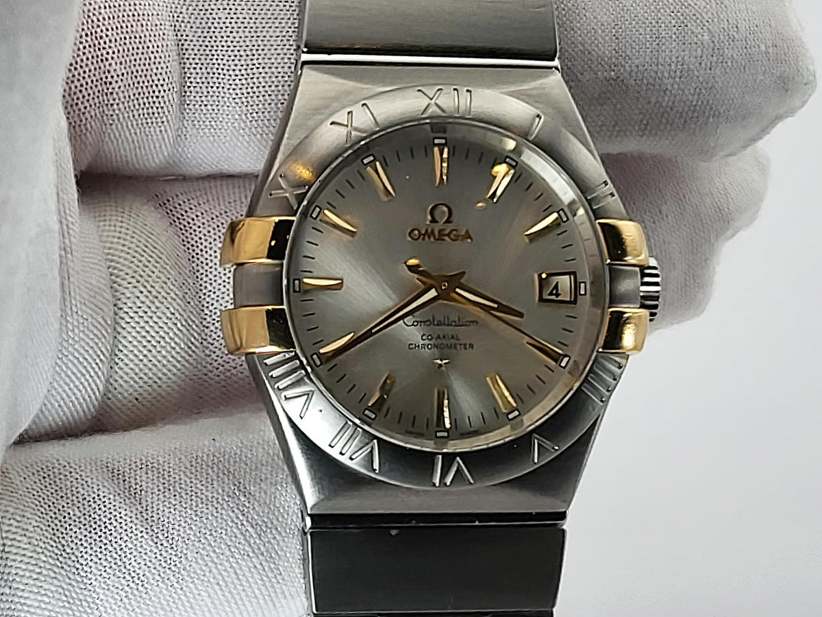 Omega Constellation wristwatch with a silver-tone stainless steel case, gold-tone accents, and a gray dial, held by a hand wearing a white textured glove.