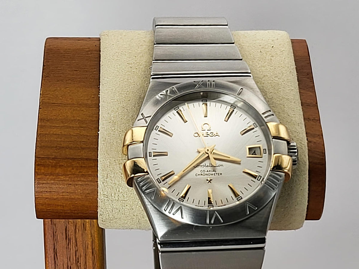 An Omega Constellation watch with a two-tone silver and gold-tone stainless steel case, a silver dial with gold-tone markers, and a silver integrated metal bracelet, displayed on a beige cushion inside a wooden stand against a light gray background.