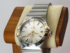 An Omega Constellation watch with a silver-tone metal bracelet and case, featuring gold-tone accents on the bezel, lugs, and dial markers, and a white dial, displayed on a wooden stand with a beige cushion.