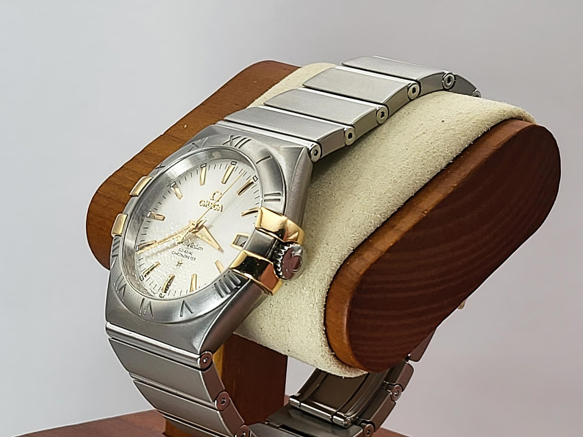 A Omega Constellation watch with a silver-tone stainless steel case and integrated bracelet, featuring a white dial with gold-tone details, displayed on a wooden stand with a cream cushion.