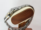 A silver-toned metal link Omega Constellation watch band with a gold-tone accent on the lugs, displayed on a wooden stand with a cream-colored cushion against a light gray background.