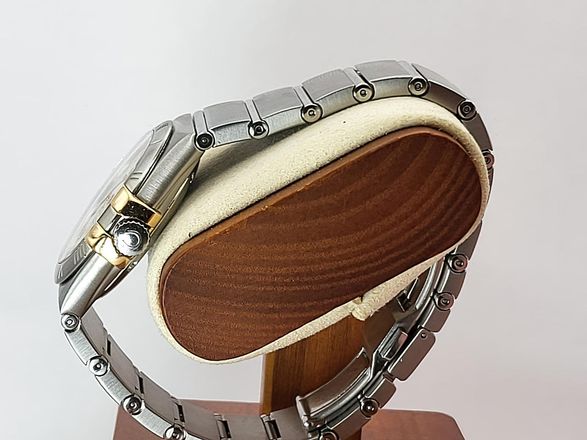 A silver-toned metal link Omega Constellation watch band with a gold-tone accent on the lugs, displayed on a wooden stand with a cream-colored cushion against a light gray background.