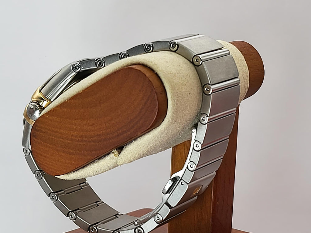 A silver-toned metal link bracelet Omega Constellation watch displayed on a wooden stand with a beige velvet cushion, photographed against a light gray background.