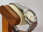 An Omega Constellation wristwatch with a two-tone silver and gold-tone metal bracelet and a cream-colored dial, displayed on a wooden stand with a beige cushion against a neutral gray background.