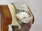 Omega Constellation watch with a two-tone stainless steel and gold-tone case, featuring a white dial with gold-tone indices and a date window, displayed on a wooden stand with a cream cushion.