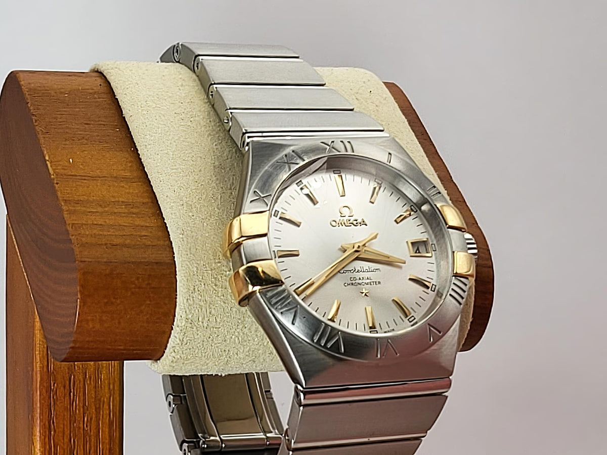 Omega Constellation watch with a two-tone stainless steel and gold-tone case, featuring a white dial with gold-tone indices and a date window, displayed on a wooden stand with a cream cushion.