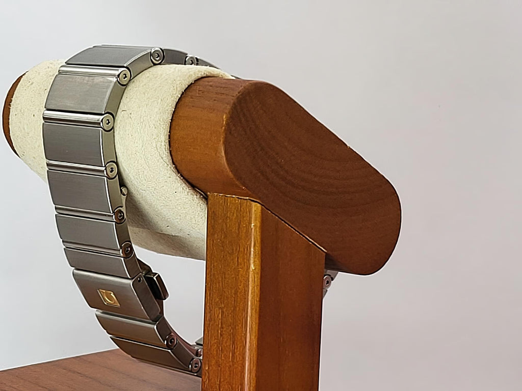 A close-up of a silver-toned metal link bracelet for a Omega Constellation watch displayed on a cream-colored cylindrical stand attached to a wooden holder, against a plain light gray background.