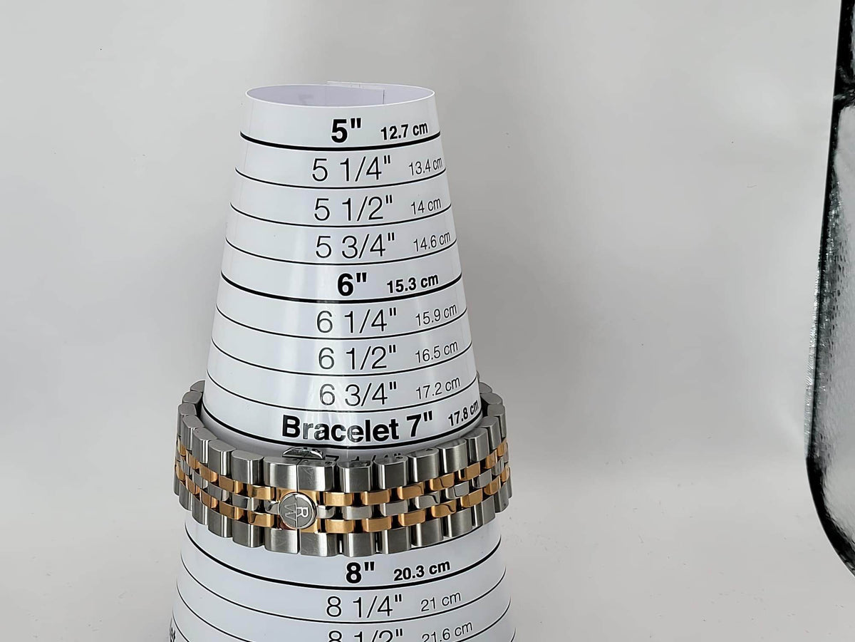 A metal bracelet for size cone stand with printed measurements in inches and centimeters, holding a silver-toned link bracelet with a gold-toned accent band for a Raymond Weil wristwatch.