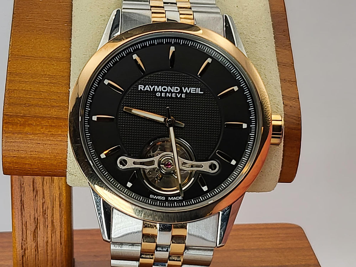 A Raymond Weil Geneve watch with a black dial, gold-tone case, and two-tone silver and gold link bracelet, displayed on a wooden stand.