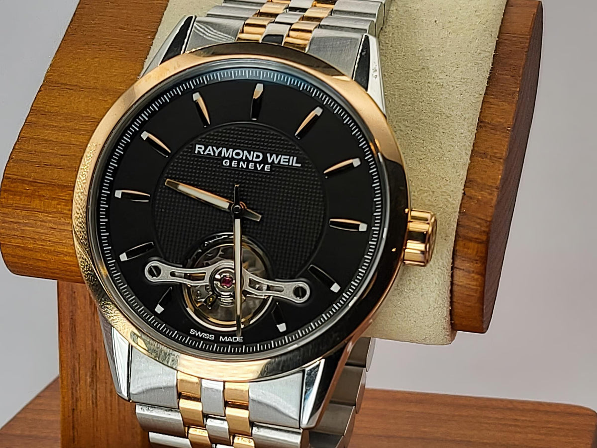 A Raymond Weil Geneve watch with a black dial, gold-tone and silver-tone two-tone metal case and bracelet, displayed on a wooden stand with a beige cushion.