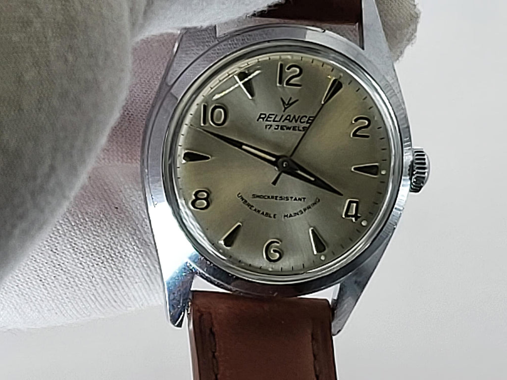 A vintage-style Reliance wristwatch with a silver-toned metal case, olive-green dial, black numerals, and a brown leather strap, photographed close up against a light neutral background with a soft fabric prop.