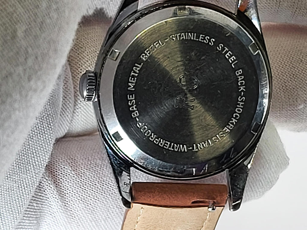Close-up of the back of a Reliance wristwatch with a brushed stainless steel case, engraved text, and a light brown leather strap resting on a white textured fabric background.