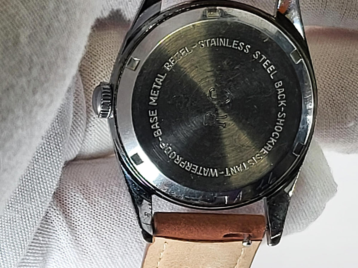Close-up of the back of a Reliance wristwatch with a brushed stainless steel case, engraved text, and a light brown leather strap resting on a white textured fabric background.