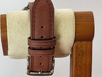 A close-up of a brown leather watch strap with silver-tone metal hardware for a Reliance wristwatch, hanging from a wooden stand with a cream-colored felt roll.