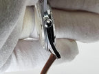 Close-up side view of a Reliance wristwatch held between white gloved hands, showing the silver-toned case, crown, and a brown strap.