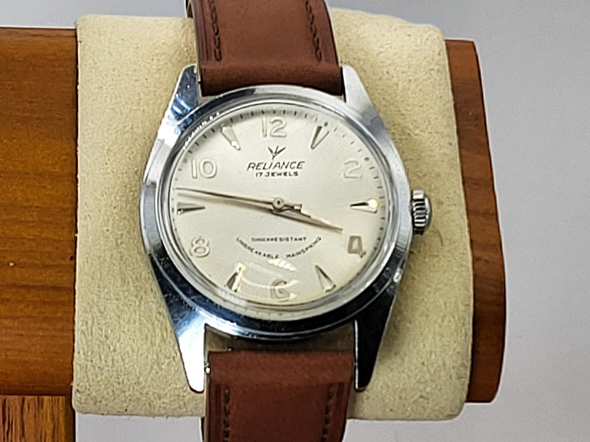 A vintage-style Reliance wristwatch with a silver-tone case, cream dial, and brown leather strap, displayed on a beige cushion mounted on a wooden stand.