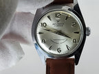 A vintage Reliance wristwatch with a silver dial and black Arabic numerals, held by a white gloved hand. The watch has a silver-tone case and a brown leather strap.