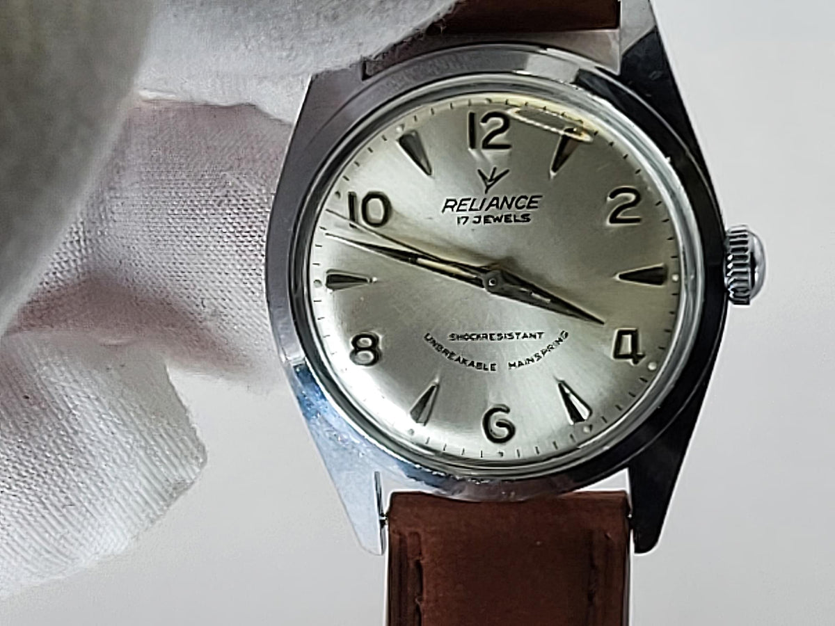 A vintage Reliance wristwatch with a silver dial and black Arabic numerals, held by a white gloved hand. The watch has a silver-tone case and a brown leather strap.