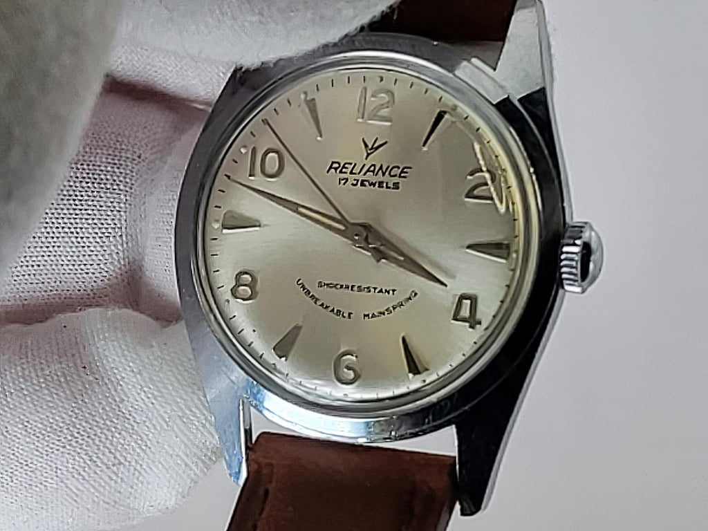 A vintage-style Reliance wristwatch with a silver-toned metal case, cream-colored dial, and brown leather strap, held against a white textured background. The dial is branded 'RELIANCE'.