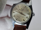 A vintage-style Reliance wristwatch with a silver-toned metal case, cream-colored dial, and brown leather strap, held against a white textured background. The dial is branded 'RELIANCE'.
