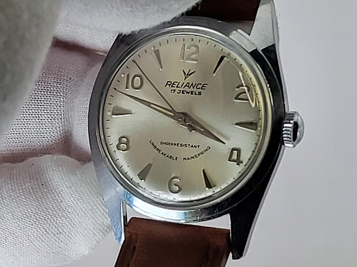 A vintage-style Reliance wristwatch with a silver-toned metal case, cream-colored dial, and brown leather strap, held against a white textured background. The dial is branded 'RELIANCE'.