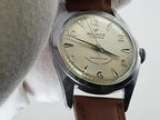 A vintage Reliance wristwatch with a silver-toned metal case, cream-colored dial, and brown leather strap, resting on a white textured surface.