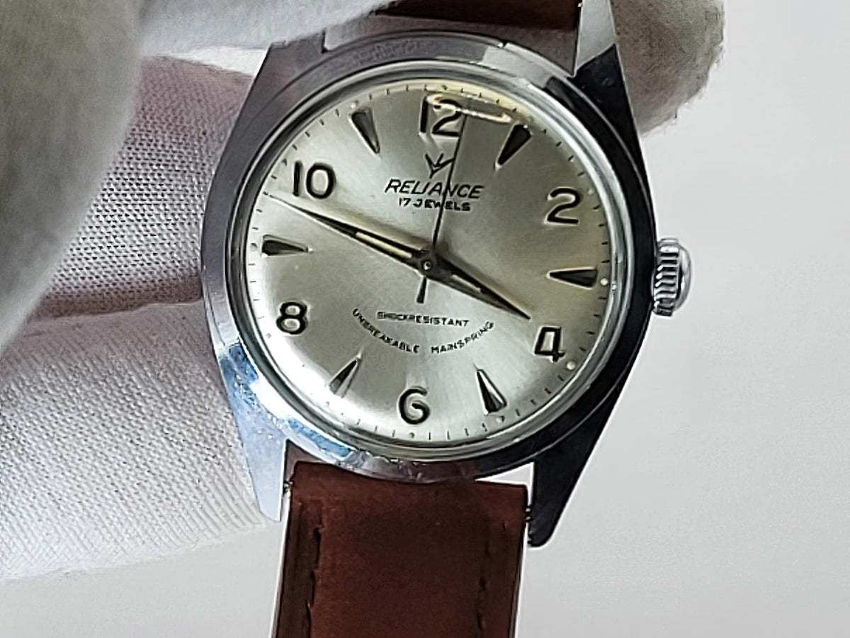 A vintage Reliance 17 jewels watch with a silver dial, black Arabic numerals, and a brown leather strap, held by a white gloved hand.
