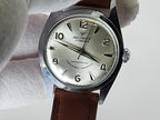 A vintage-style Reliance wristwatch with a silver-toned metal case, a cream-colored dial featuring black Arabic numerals and hands, and a brown leather strap. The watch is held by a white gloved hand against a plain light background.