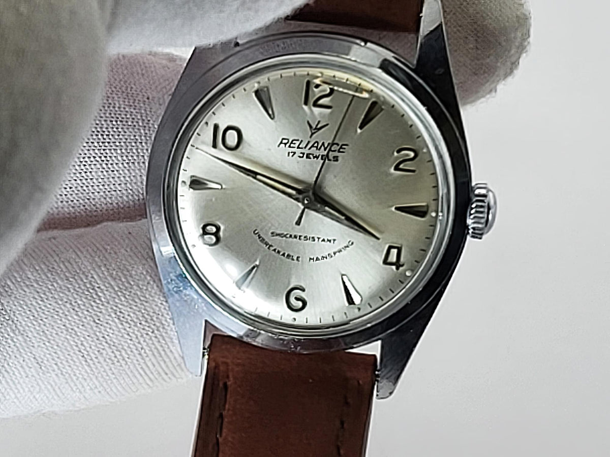 A vintage-style Reliance wristwatch with a silver-toned metal case, a cream-colored dial featuring black Arabic numerals and hands, and a brown leather strap. The watch is held by a white gloved hand against a plain light background.