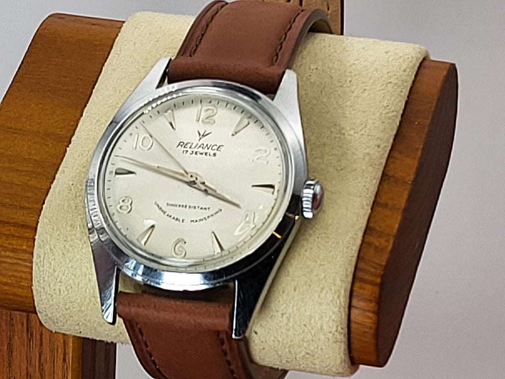 A vintage-style Reliance wristwatch with a brown leather strap and a silver-toned case, displayed on a beige cushion inside a wooden stand. The watch face is cream-colored with black Arabic numerals and the brand name 'Reliance' is visible.
