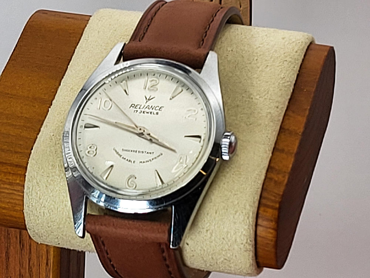 A vintage-style Reliance wristwatch with a brown leather strap and a silver-toned case, displayed on a beige cushion inside a wooden stand. The watch face is cream-colored with black Arabic numerals and the brand name 'Reliance' is visible.
