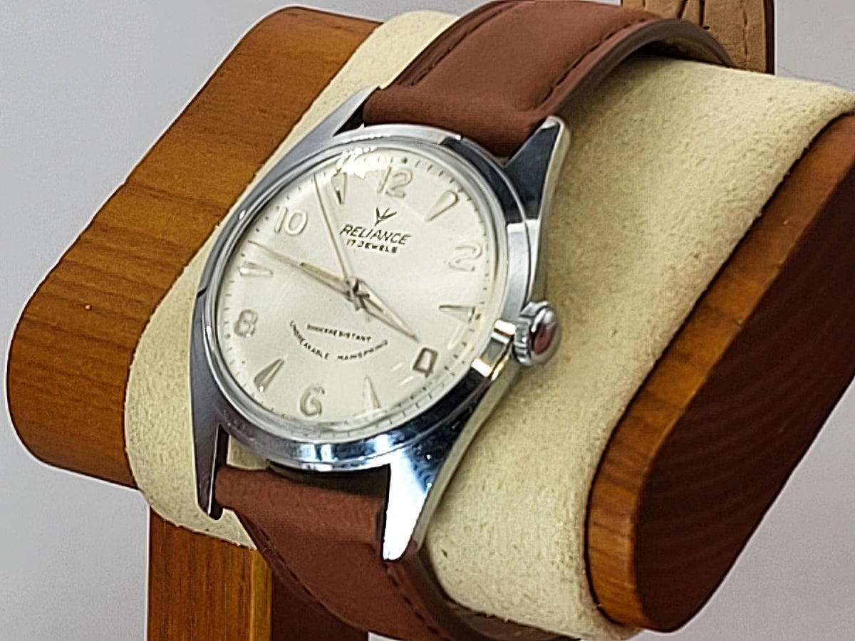 A vintage-style Reliance wristwatch with a brown leather strap, silver-toned metal case, and cream-colored dial, displayed on a wooden stand with a beige cushion.