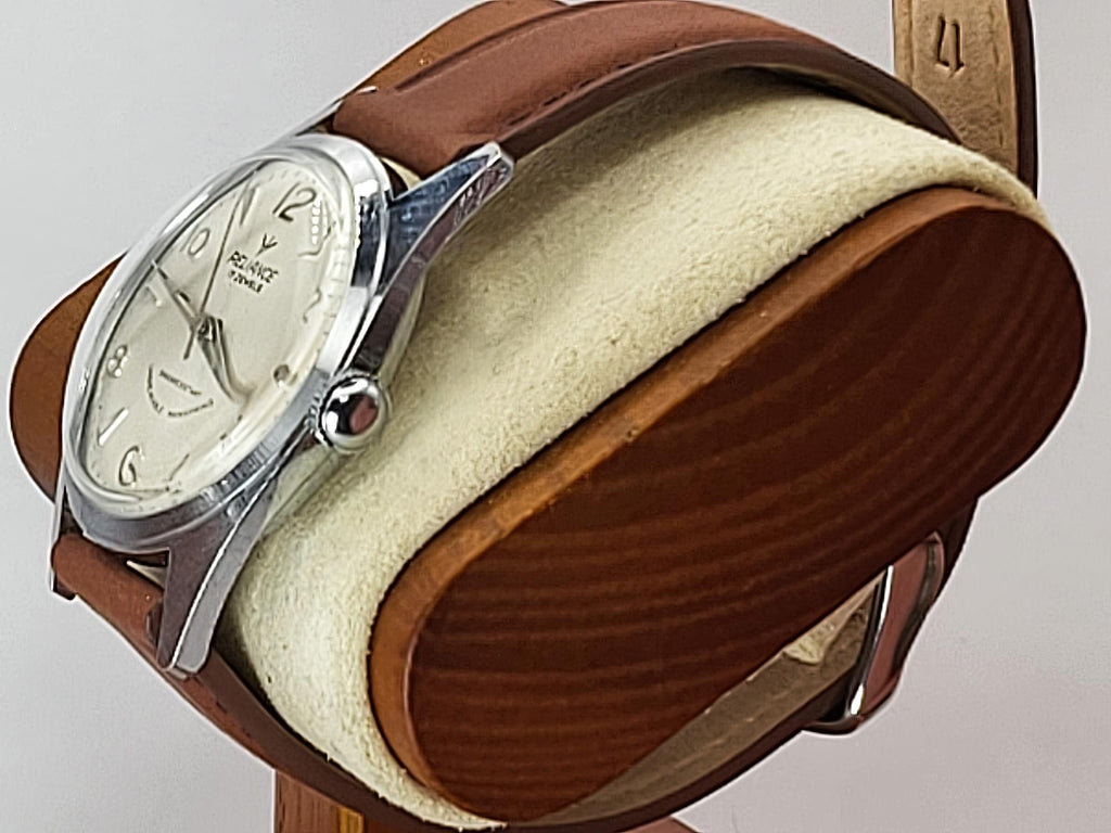 A vintage Reliance wristwatch with a silver-tone case, cream dial, and brown leather strap, displayed on a wooden and felt watch stand against a light gray background.