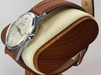 A vintage Reliance wristwatch with a silver-tone case, cream dial, and brown leather strap, displayed on a wooden and felt watch stand against a light gray background.