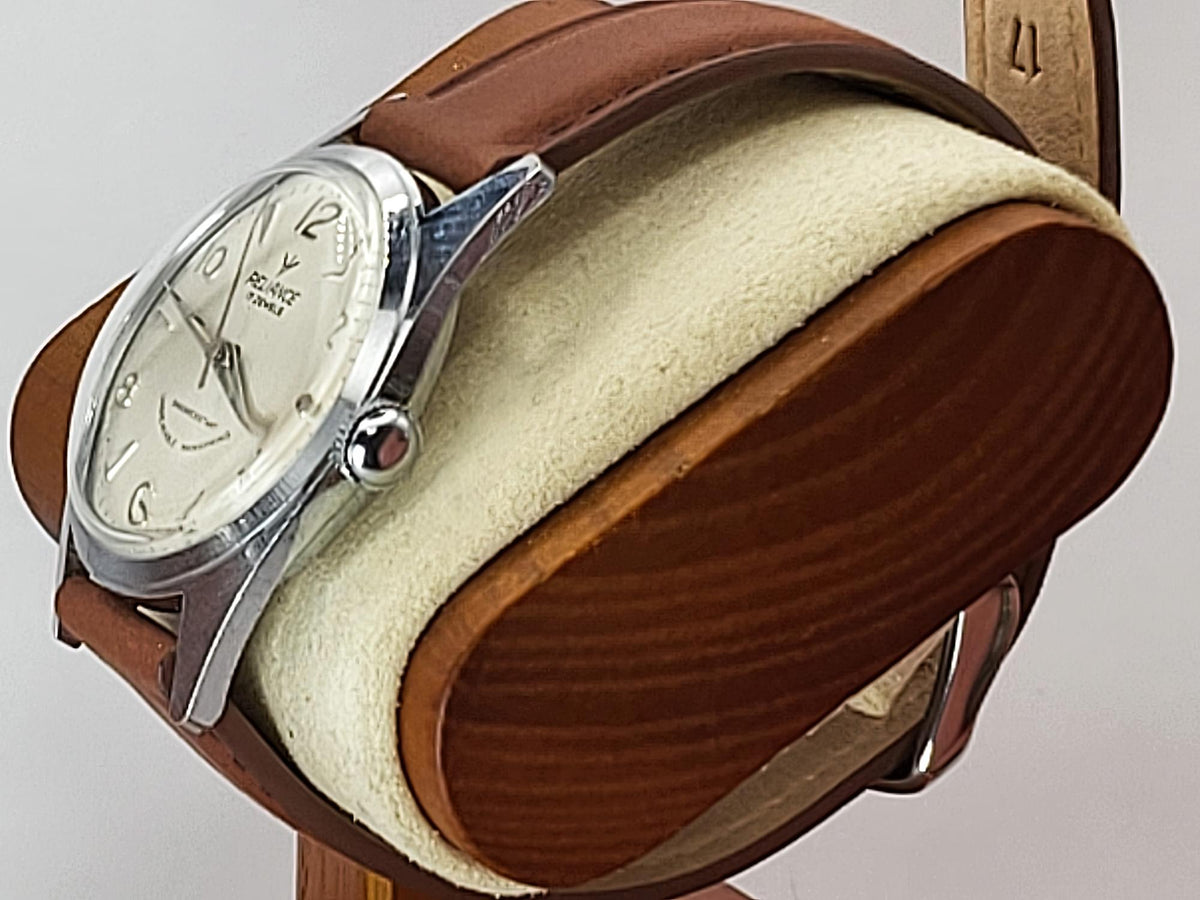 A vintage Reliance wristwatch with a silver-tone case, cream dial, and brown leather strap, displayed on a wooden and felt watch stand against a light gray background.