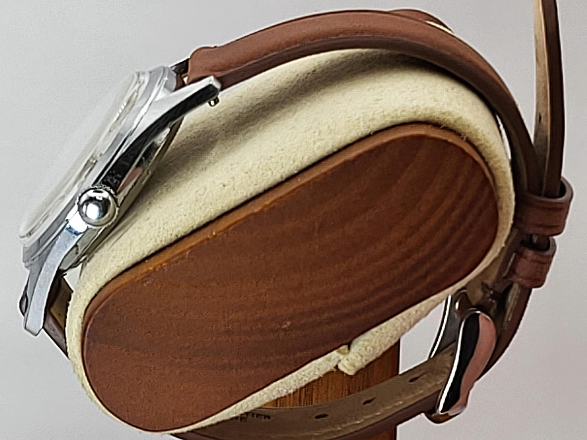 A close-up of a Reliance wristwatch with a brown leather strap and silver-toned metal case, resting on a round wooden display stand with a cream felt lining.