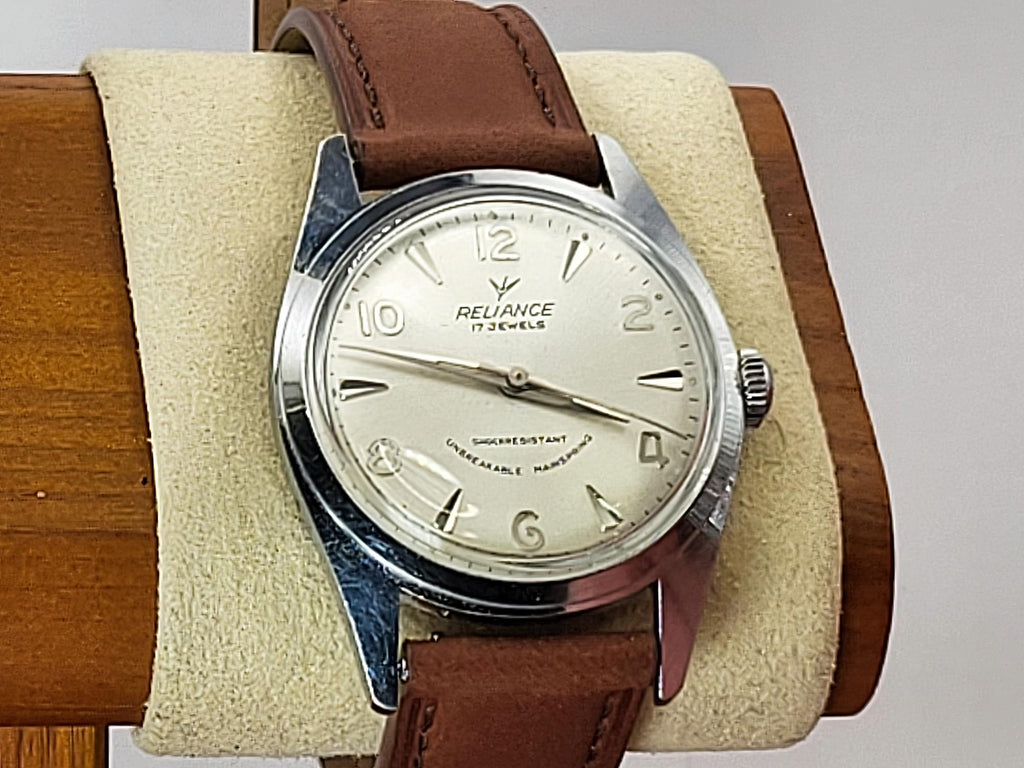 A Reliance vintage-style wristwatch with a silver-tone round case, cream dial, and brown leather strap, displayed on a beige cushion mounted on a wooden stand.