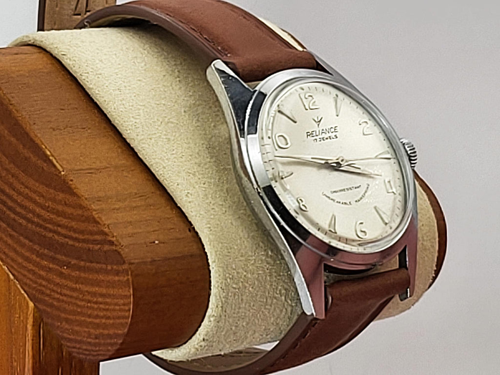 A vintage-style Reliance wristwatch with a brown leather strap, silver-toned metal case, and cream-colored dial, displayed on a beige cushion mounted on a wooden stand against a light gray background.