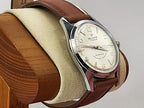 A vintage-style Reliance wristwatch with a brown leather strap, silver-toned metal case, and cream-colored dial, displayed on a beige cushion mounted on a wooden stand against a light gray background.
