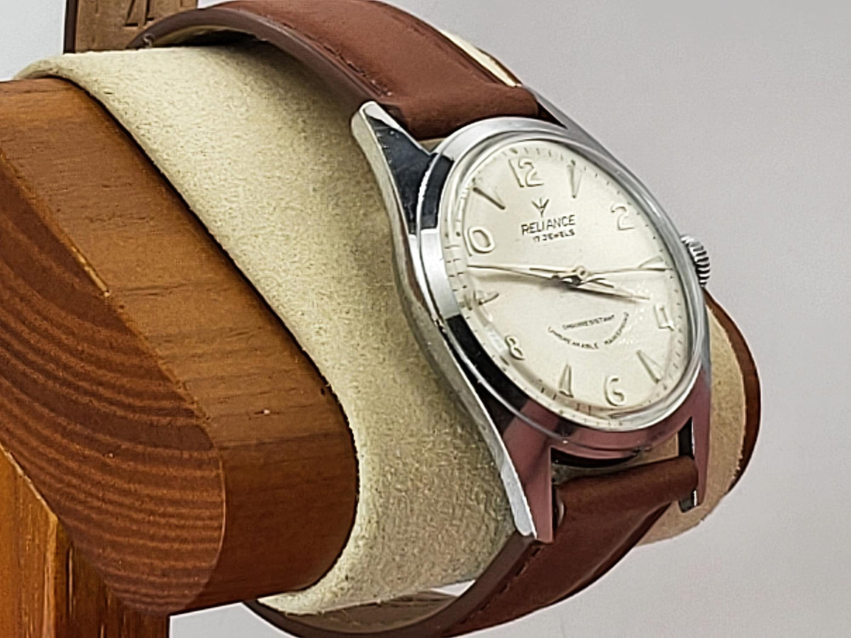 A vintage-style Reliance wristwatch with a brown leather strap, silver-toned metal case, and cream-colored dial, displayed on a beige cushion mounted on a wooden stand against a light gray background.