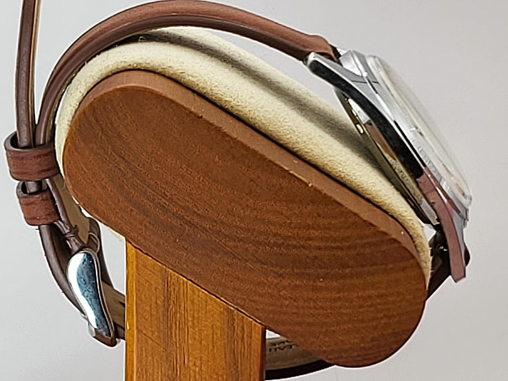 A close-up of a Reliance wristwatch with a brown leather strap and silver-toned case, resting on a wooden display stand with a cream cushion.