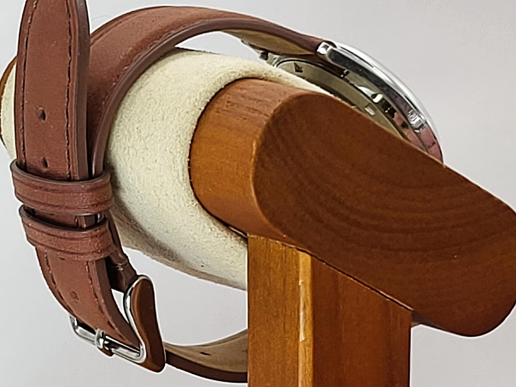 A close-up of a brown leather watch strap with a silver buckle for a Reliance wristwatch, draped over a wooden and cream-colored watch display stand.