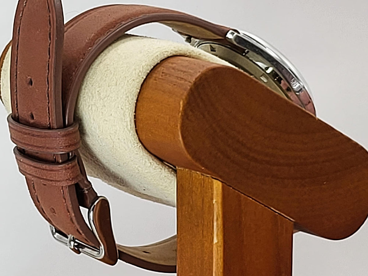 A close-up of a brown leather watch strap with a silver buckle for a Reliance wristwatch, draped over a wooden and cream-colored watch display stand.